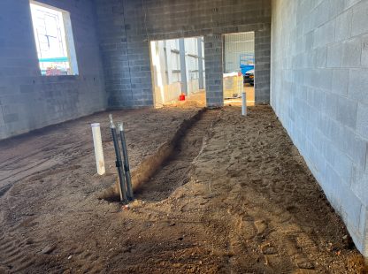 Interior of a concrete block room with a dirt floor, showing a narrow trench cut through the center, vertical utility pipes, and an open doorway leading to another unfinished area.
