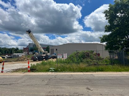 Construction site viewed from the road, with a crane lifting materials beside a metal-sided building, warning signs posted along a grassy area, and traffic cones near the entrance under cloudy skies.