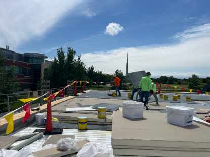 Workers installing rooftop insulation boards, with stacked materials, buckets, and safety flags across a flat roof near a modern building and trees under a blue sky.