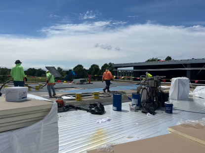 Construction workers installing insulation boards on a flat metal roof, with tools, buckets, and materials spread across the surface and a modern building in the background under a cloudy sky.