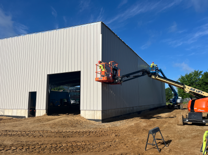 Workers on boom lifts installing vertical metal siding on a large building exterior, with dirt grading and a wide open doorway at the construction site under a clear blue sky. Provide your feedback on BizChat