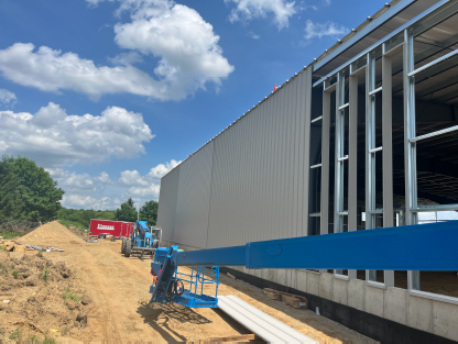 Partially enclosed steel building with vertical siding installed on one wall and exposed framing on the other, with a boom lift, construction materials, and a dirt work path alongside the site under a blue sky with scattered clouds.