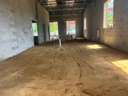 Interior of a concrete block building under construction with exposed steel roof trusses, sandy floor with tire marks, utility pipes protruding, and window and door openings letting in daylight.