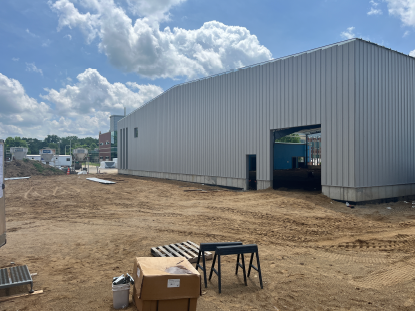 Large metal-sided building with curved roofline and open doorway, surrounded by a graded dirt construction site with materials and equipment in the foreground under a partly cloudy sky.