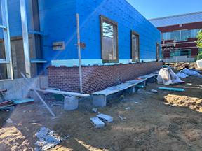 Brick veneer foundation wall beneath a blue sheathed exterior, with window openings, construction materials, and scaffolding along a dirt worksite next to a larger building.