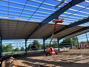 Interior of a steel-framed building with partial roof panels installed, featuring a boom lift raised to the ceiling over a sand-covered floor.