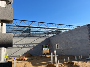 Interior of a steel-framed building with partial roof panels installed, featuring a boom lift raised to the ceiling over a sand-covered floor.