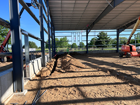 Interior of a steel-framed building with roof panels installed, showing a sand-covered floor, long shadows from the structure, and construction equipment parked along the perimeter.
