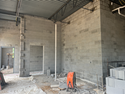 Interior corner of a concrete block building under construction, with exposed steel roof decking overhead, a temporary support pipe, stacked masonry blocks, and construction materials scattered across the unfinished floor.