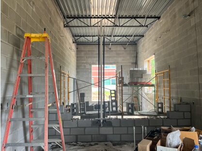 Interior of a concrete block building under construction, featuring a partially built masonry partition wall, scaffolding, a tall ladder, exposed steel roof decking, and construction materials throughout the unfinished space.