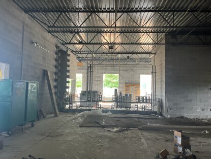 Interior of a large concrete block building under construction, featuring exposed steel roof trusses and metal decking, partial masonry partition walls, scaffolding, and an unfinished concrete floor with construction materials throughout the open space.