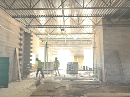 Construction workers building an interior concrete block partition wall inside a large unfinished structure, with scaffolding, exposed steel roof trusses and metal decking overhead, and construction materials spread across the floor.