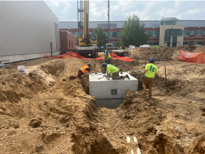 Workers installing a precast concrete utility structure within a large excavation trench beside a building under construction, with a crane, exposed utility trenches, and construction fencing visible throughout the dirt worksite.