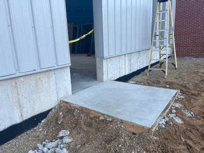 Newly poured concrete sidewalk and entrance landing at a metal building under construction, connecting the building doorway to the surrounding grade, with a ladder, exposed foundation wall, and unfinished landscaping visible around the entrance.