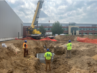 Workers installing a precast concrete utility structure within a deep excavation beside a metal building under construction, with a crane positioned overhead, utility trenches extending through the site, and safety fencing surrounding the work area.