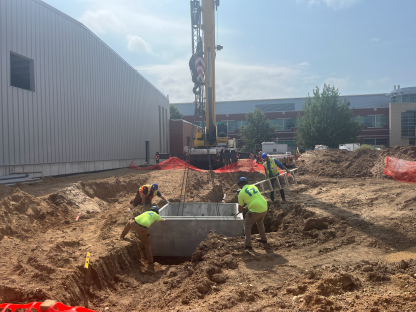 Construction crew guiding a precast concrete utility structure into a deep excavation using a crane, with workers positioned around the trench, safety fencing, and active earthwork alongside a metal building under construction.