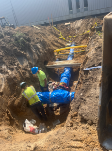 Workers installing and connecting underground utility piping within a deep excavation trench, with large blue-wrapped pipes, trench shoring, and safety barricade tape visible alongside a building construction site.