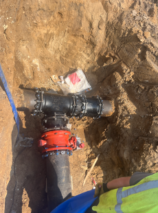 Close-up view of underground utility piping installed within an excavation trench, showing a large black pipe connected through a tee fitting with red and blue couplings, surrounded by exposed soil during site utility construction.