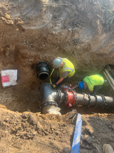 Construction workers installing large underground utility piping within a deep excavation trench, connecting pipe sections and fittings while standing beside exposed utility infrastructure in a dirt worksite adjacent to a building under construction.