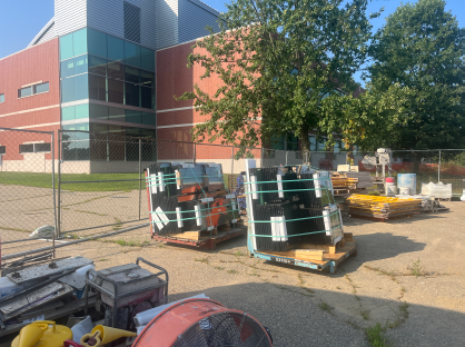 Stacks of glass panels and construction materials stored on pallets within a fenced staging area beside a brick-and-glass building, with additional equipment and supplies organized for ongoing construction work.