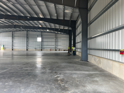 Wide interior view of a newly constructed metal building, featuring a finished polished concrete floor, corrugated metal walls, exposed steel roof framing, and workers inspecting or measuring along the perimeter wall near a large overhead door opening.