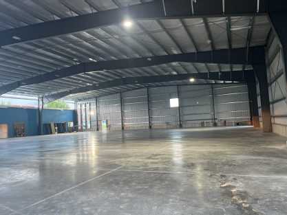 Wide interior view of a newly constructed metal building, featuring a finished polished concrete floor, corrugated metal walls, exposed steel roof framing, and workers inspecting or measuring along the perimeter wall near a large overhead door opening.