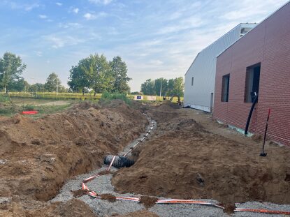 Excavated utility trench running alongside a partially completed brick and metal building, with drainage pipe, gravel bedding, and exposed conduit visible in the dirt work area under a partly cloudy sky.