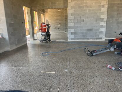 Finished polished concrete floor inside a concrete block building, with construction equipment, extension cords, and unfinished doorways leading to adjacent rooms under active interior construction.