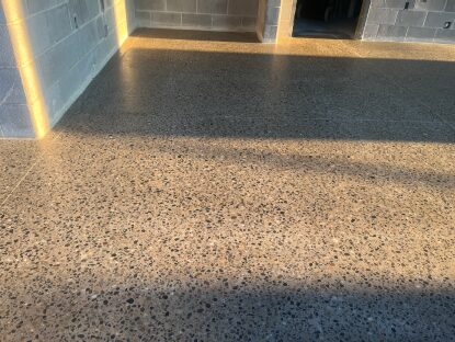 Polished exposed-aggregate concrete floor inside an unfinished concrete block building, with sunlight casting long shadows across the room and multiple door openings leading to adjacent spaces.