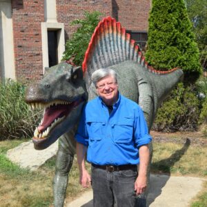Robb Gillespie wearing a blue shirt standing next to a life size 3D model of a dinosaur with sharp teeth.
